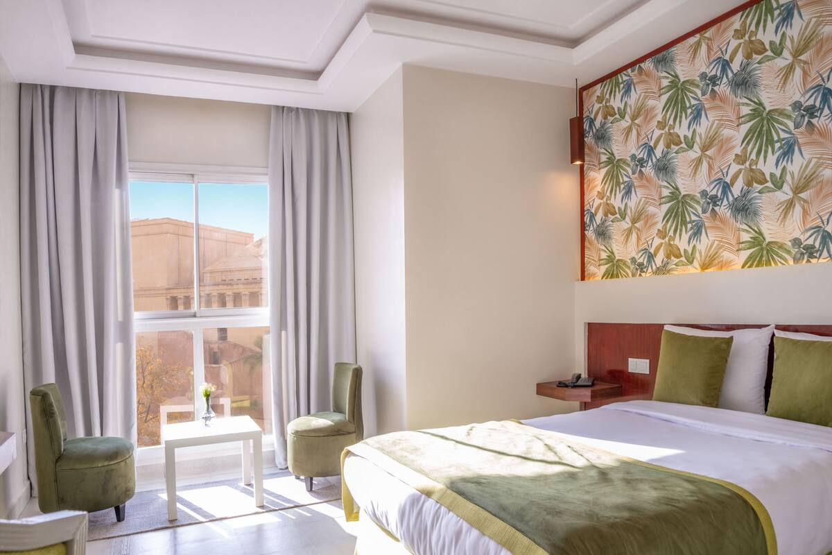 A hotel room with a large bed, a desk, two chairs, a window with curtains, and a tropical wallpaper.