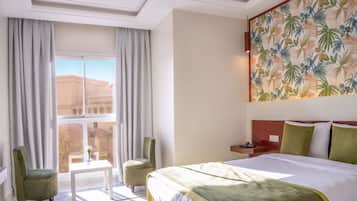 A hotel room with a large bed, a desk, two chairs, a window with curtains, and a tropical wallpaper.