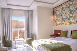 Habitación de hotel con una cama grande, un escritorio, dos sillas, una ventana con cortinas y un papel tapiz tropical.