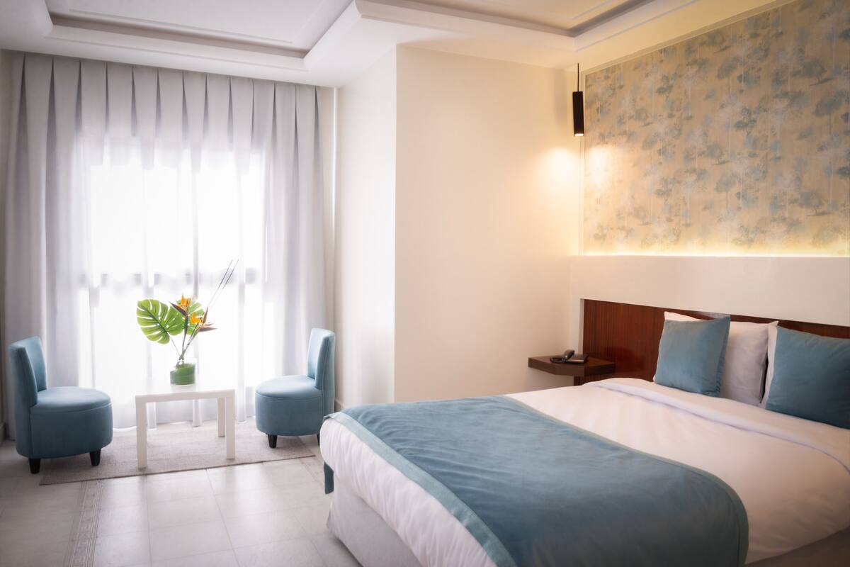 A hotel room with a large bed, two armchairs, a small table, and a potted plant.