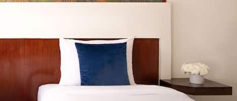 A hotel room with a wooden headboard, a white bedspread, a blue pillow, and a small nightstand with a vase of flowers.
