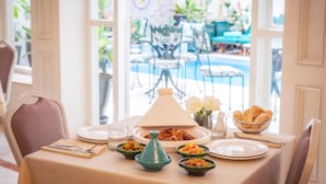 Mesa de jantar posta com pratos, tigelas e um bule, com vista para área externa com mesas e uma piscina.