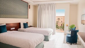 Quarto de hotel com duas camas, uma janela grande com cortinas, vista para o exterior e um espaço de estar com uma cadeira e uma planta.