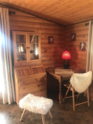 Een gezellig interieur van een chalet met houten wanden, een houten kastje met glazen deuren, een rode lamp, een witte zachte poef en een stoel.
