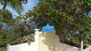 A white gate with a view of the ocean, surrounded by lush greenery and flowering plants. - Beachfront luxury modern 2-bed apt with pool by BSL Rentals (Paynes Bay)