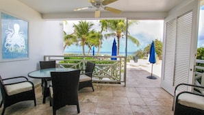 An outdoor patio with a table and chairs, a ceiling fan, and a view of palm trees and the ocean. - Beachfront luxury modern 2-bed apt with pool by BSL Rentals (Paynes Bay)