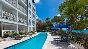 A swimming pool with lounge chairs and umbrellas, surrounded by a multi-story building with balconies. - Beachfront luxury modern 2-bed apt with pool by BSL Rentals (Paynes Bay)