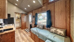 Una habitación compacta tipo cabaña con paredes y techo de madera, una kitchenette, una cama con vistas y una ventana con cortinas.