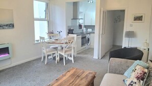 A living room with a sofa, a wooden coffee table, a dining table with chairs, a kitchen area with a stove and washing machine, and a fireplace. - Angel Lane Apartment. Alnwick Town Centre (Alnwick)