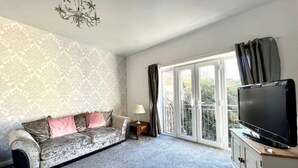 A living room with a sofa, a television on a cabinet, a chandelier, and a large window with curtains. - Angel Lane Apartment. Alnwick Town Centre (Alnwick)