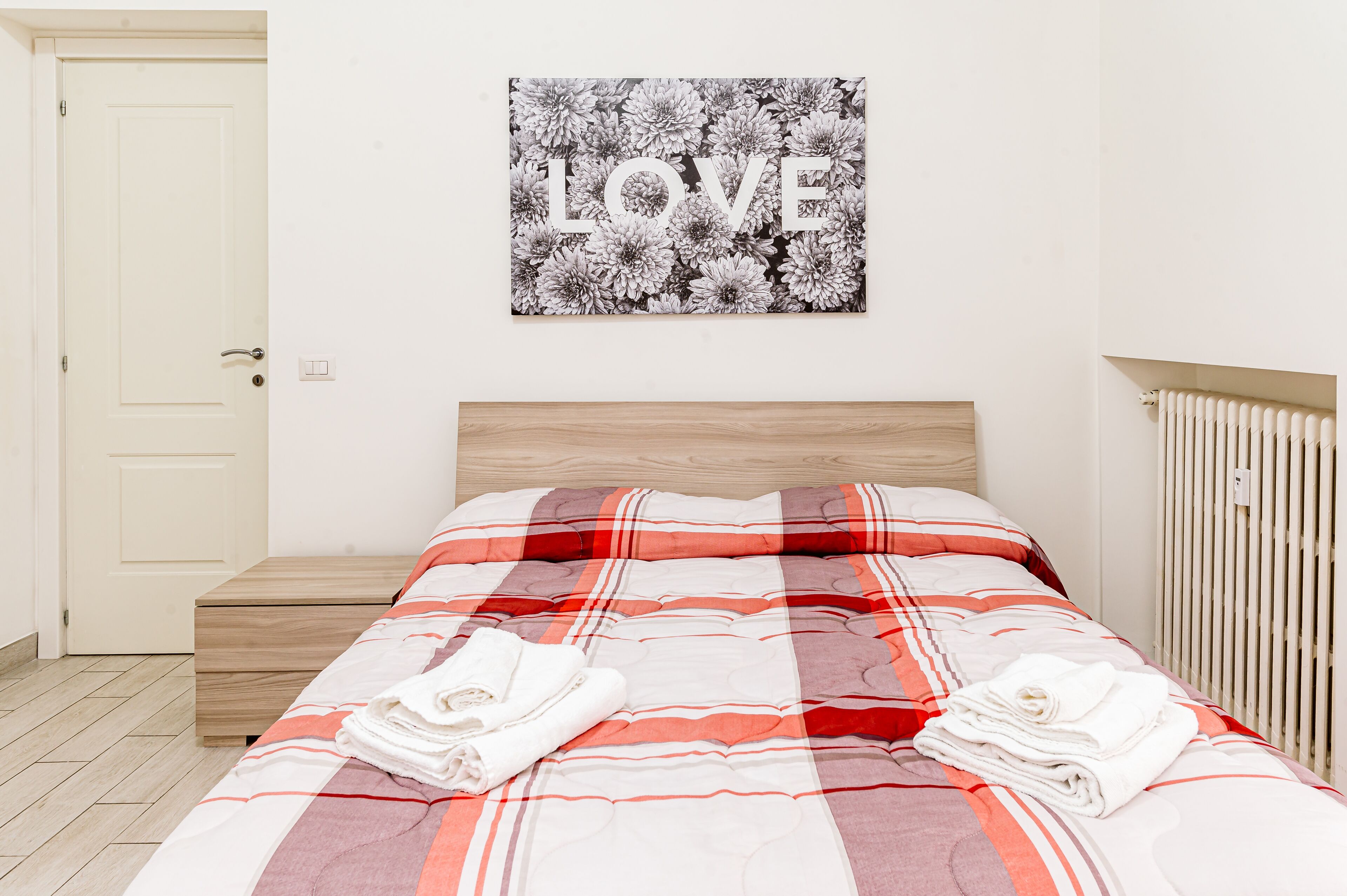 A neatly made bed with a red and white striped blanket, a wooden headboard, and a wall-mounted picture of flowers with the word "LOVE".