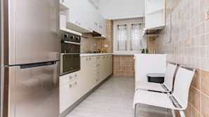 Uma cozinha compacta com geladeira de aço inoxidável, forno embutido e pia. Há uma pequena área de refeições com uma cadeira e uma mesa.