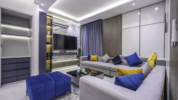 A modern living room with a grey sofa, blue and yellow cushions, a flat-screen TV, and a built-in air conditioning unit. - Mabaat - Al Aqiq - 504 (Riyadh)