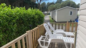 Une terrasse en bois avec des chaises longues en plastique blanc, une clôture en bois et, au fond, une rangée de maisons mobiles.