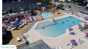 Une piscine entourée de chaises longues et de parasols, elle-même délimitée par une clôture, avec d’autres chaises longues à proximité et un bâtiment visible au loin.