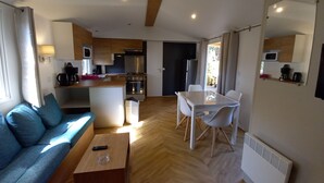 Un salon moderne avec un canapé, une table basse et un coin repas avec des chaises. On aperçoit une cuisine à l’arrière-plan, équipée d’appareils électroménagers et d’un four à micro-ondes.