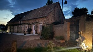 Un bâtiment rustique en pierre, doté d’un toit en pente, d’un petit porche et d’une statue placée au premier plan.