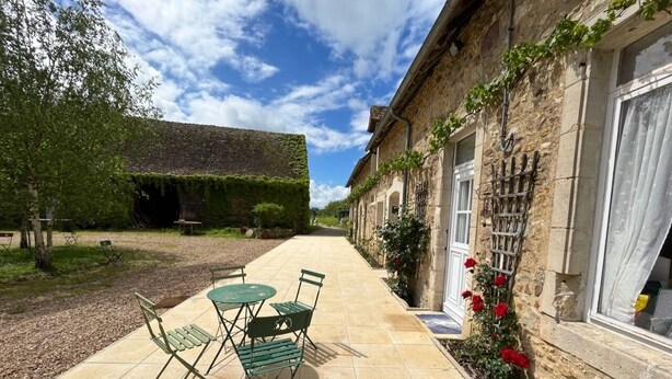Une cour avec une table et des chaises, un bâtiment en pierre doté de fenêtres et un arbre.