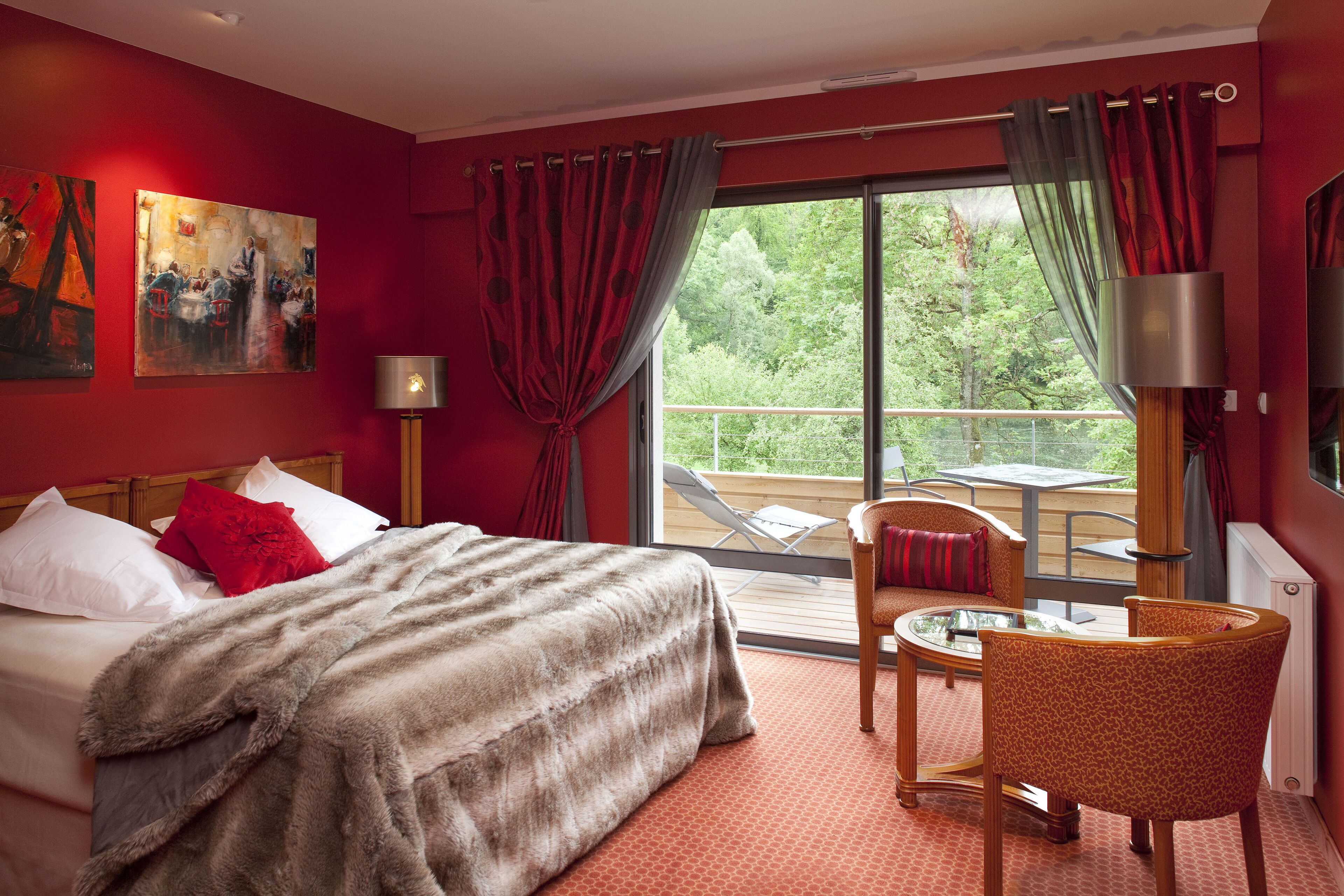 A hotel room with a bed, a desk, a chair, a window with a view of greenery, and artwork on the wall.