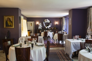 Comedor con mesas redondas, sillas y manteles blancos. Hay lámparas fijadas a la pared y un espejo decorativo.