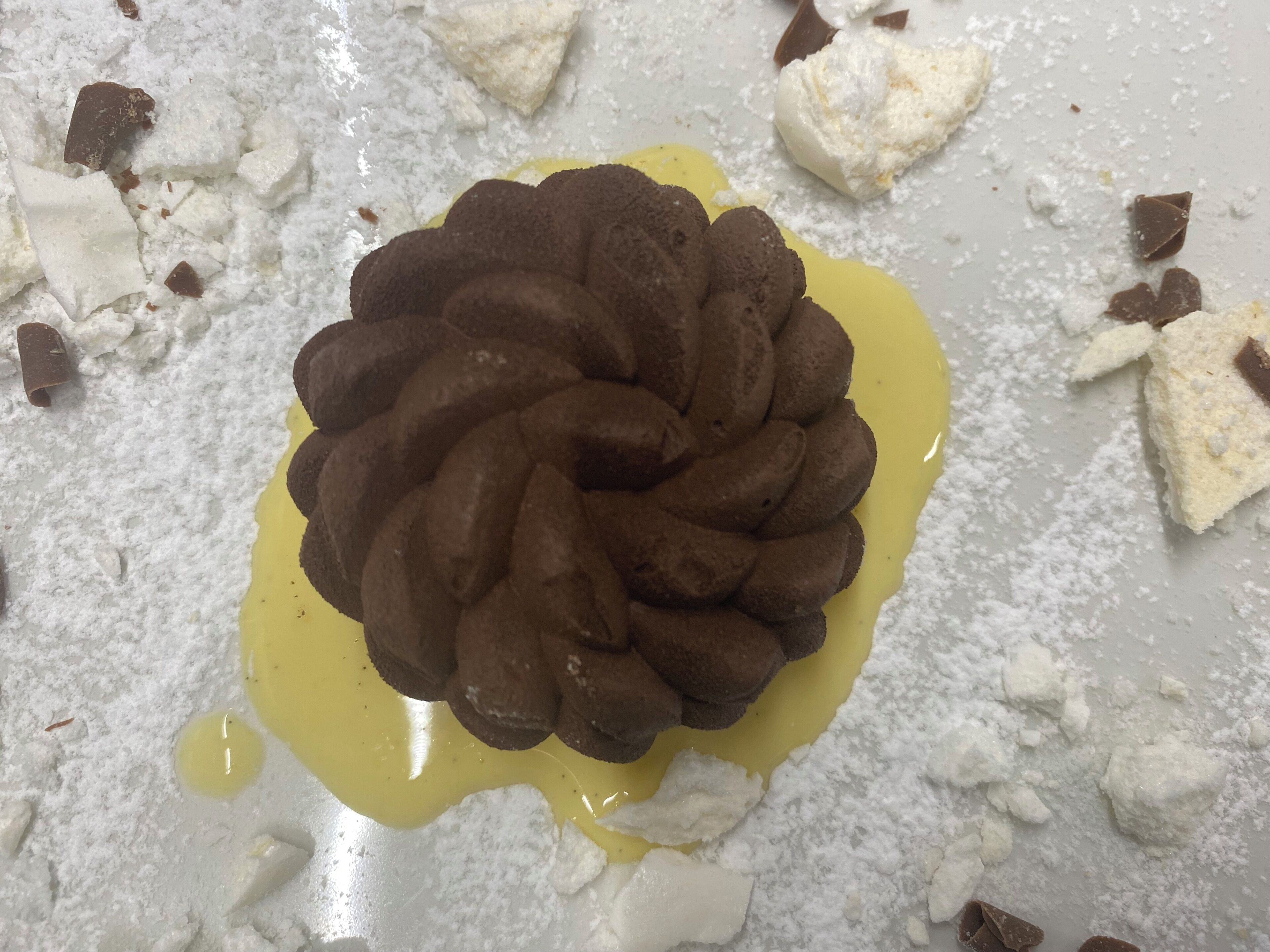 Un postre con base de chocolate y salsa amarilla, rodeado de azúcar glas y trozos de chocolate.