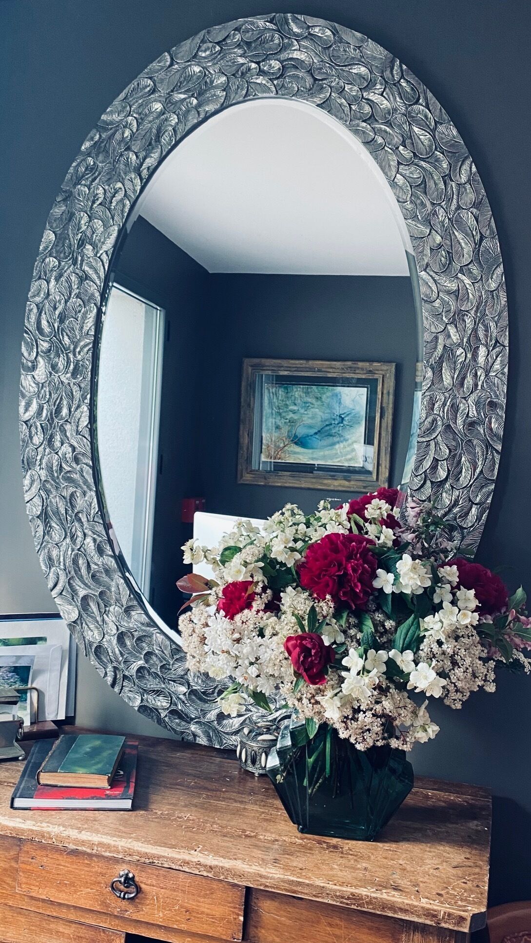 A mirror with a leaf pattern, a vase of flowers, and a wooden dresser with books.