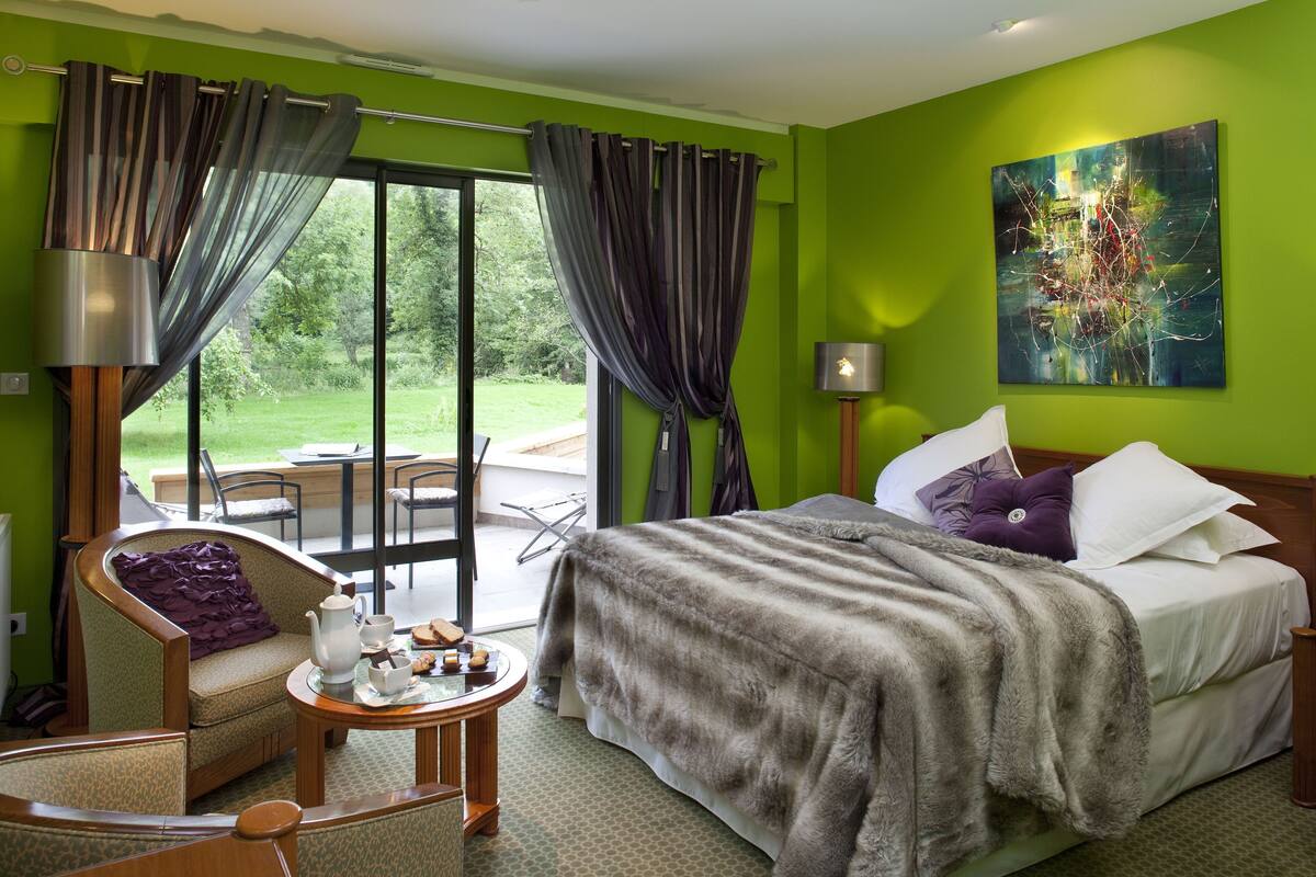 A hotel room with a bed, a chair, a table with tea set, a painting on the wall, and a view of the outdoors.