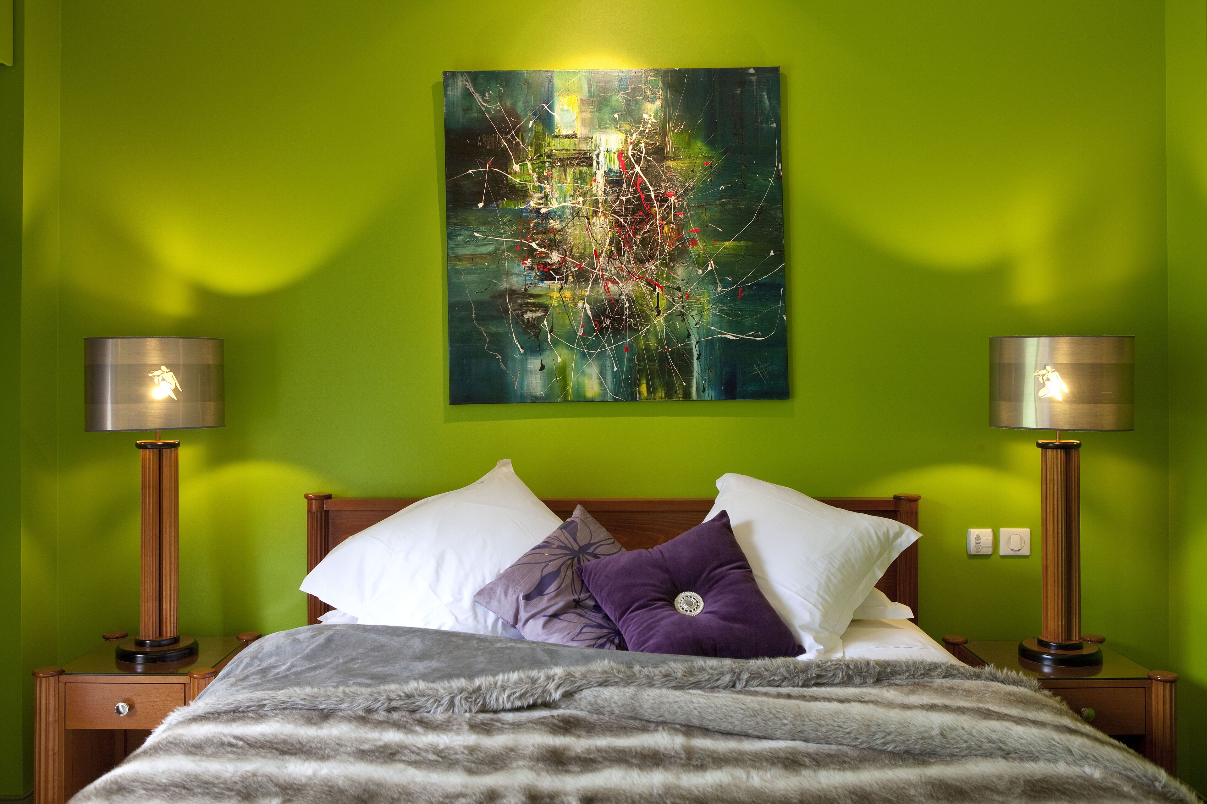 A neatly made bed with a gray blanket, two bedside tables with lamps, and a green wall with a large abstract painting.