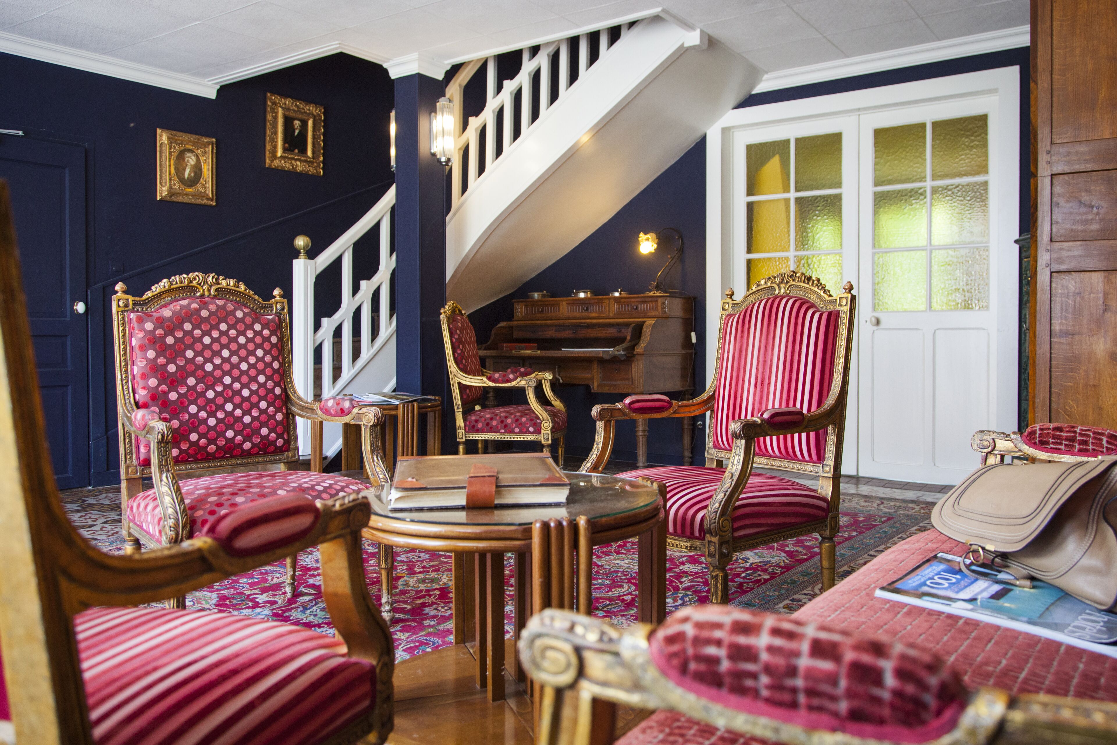 A room with a staircase, ornate chairs, a piano, and a patterned rug.