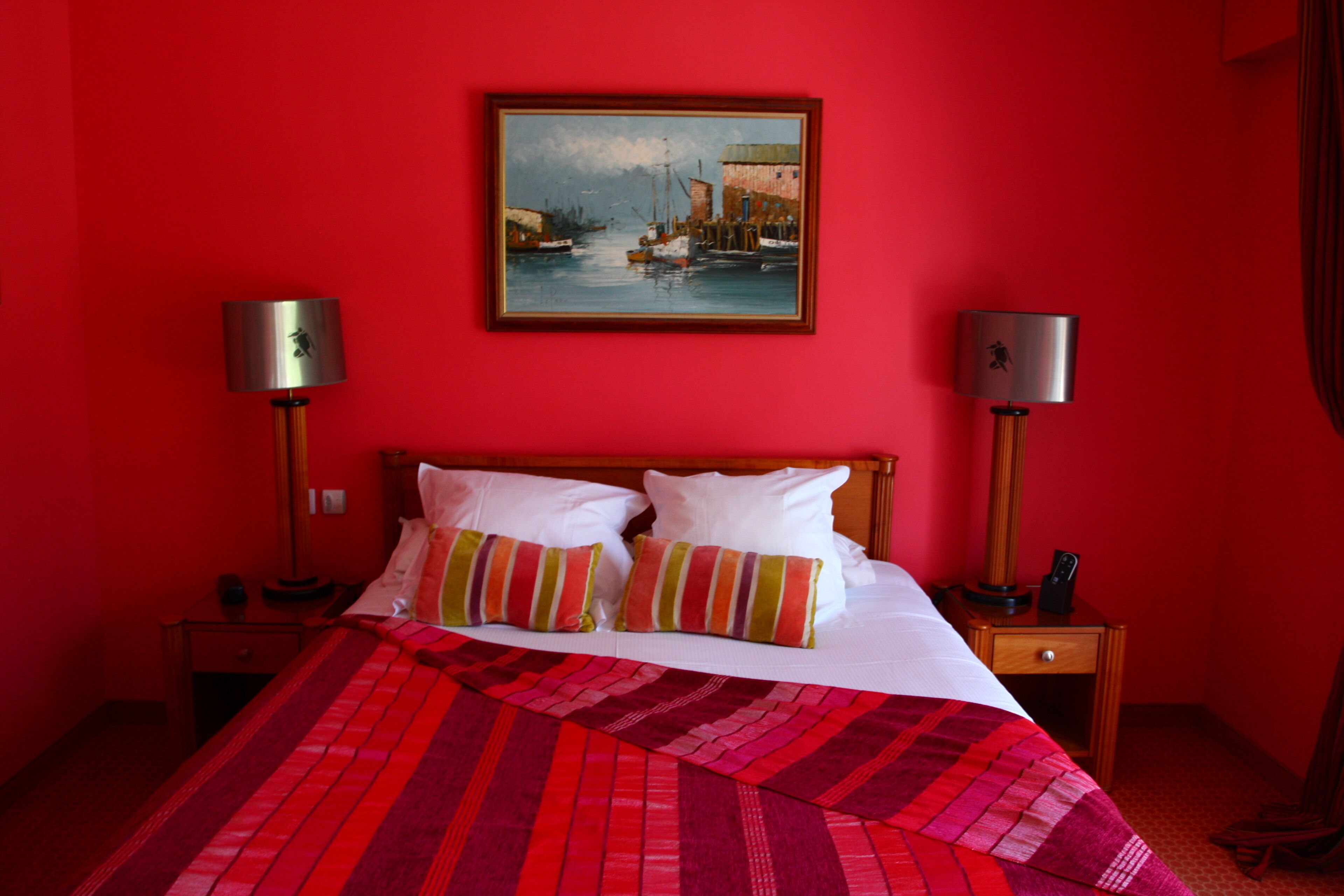Ein Hotelzimmer mit roter Wand, einem Bett mit weißem und gestreiftem Kissen, einer rot-schwarz gestreiften Decke, zwei Nachttischlampen und einem Bild von Booten an der Wand.