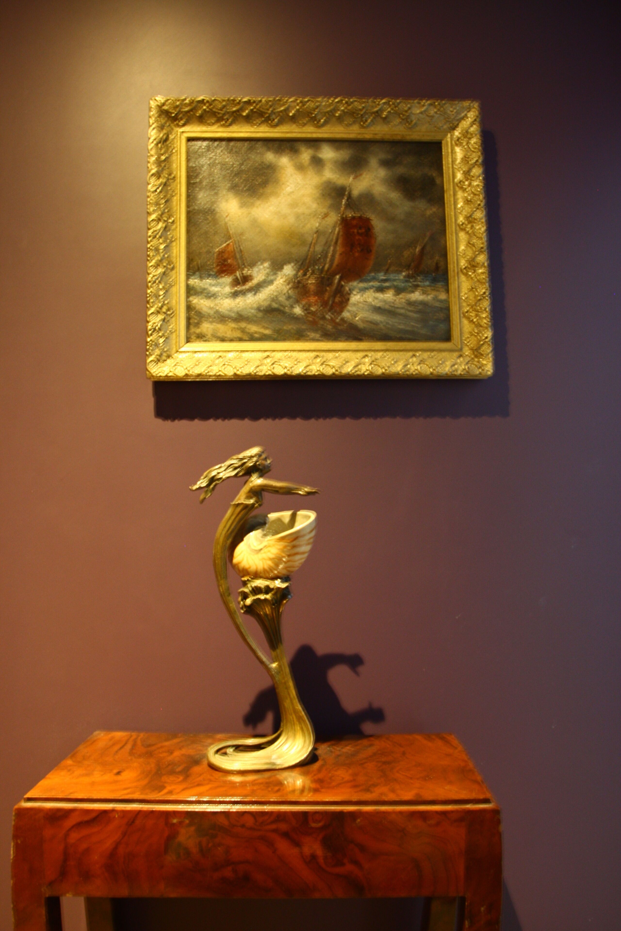 A framed painting of a ship on the sea, mounted on a wall. Below it, there is a decorative metal sculpture on a wooden table.