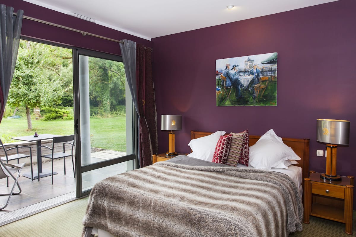 A bedroom with a large bed, two bedside tables with lamps, a painting on the wall, and a view of the outdoors through a sliding door.