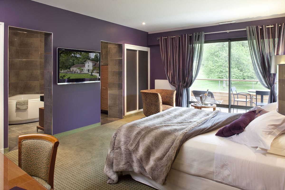 A modern hotel room with a large bed, a television, a bathroom, and a balcony with a view of the outdoors.