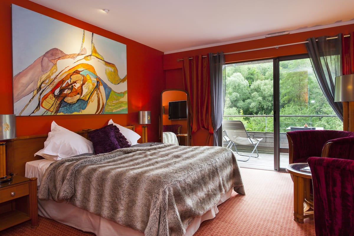 A hotel room with a large bed, a painting on the wall, a balcony with a chair, and a view of greenery.