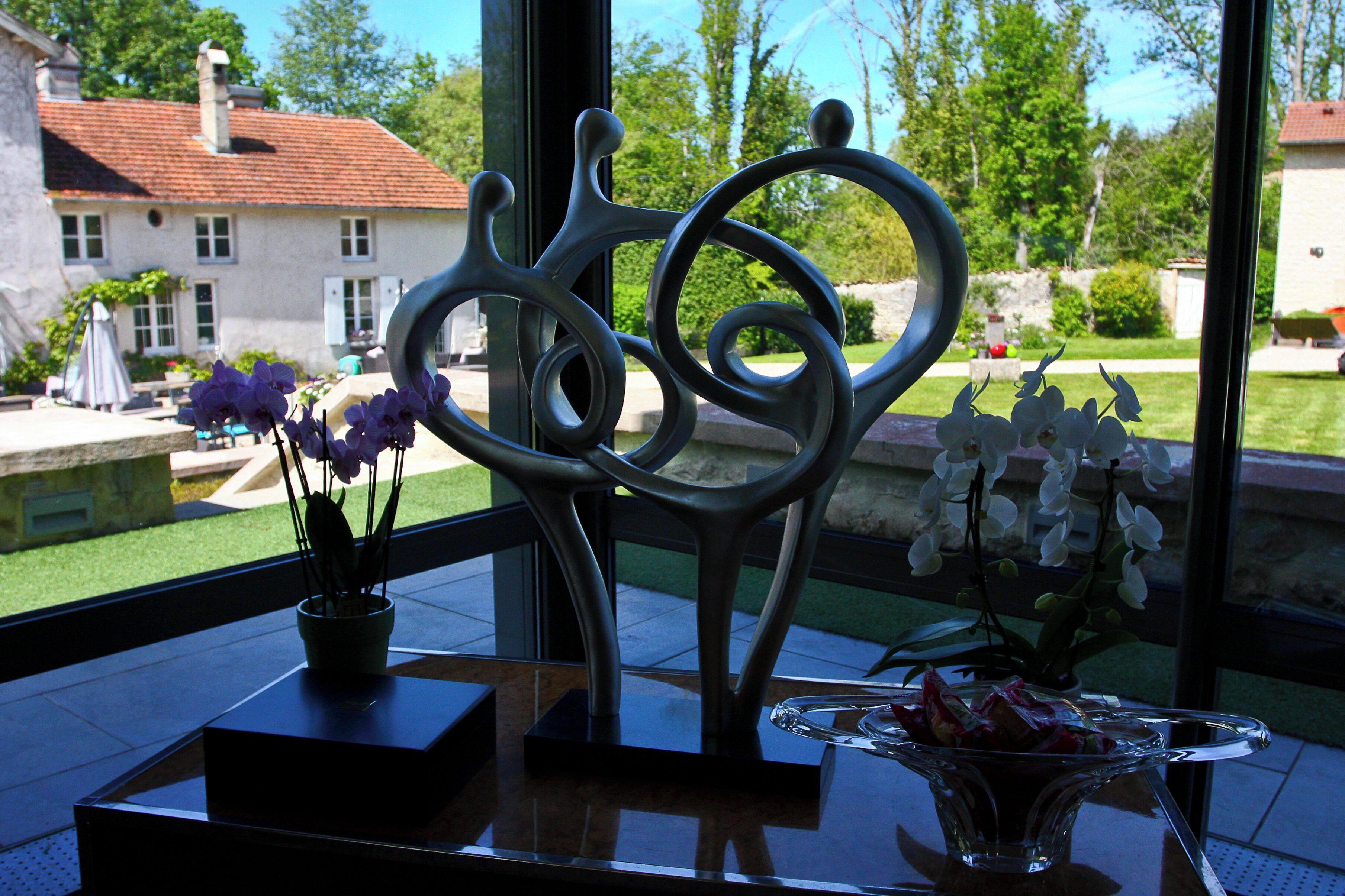 A room with a view of a garden, a metal sculpture, and two flower arrangements on a table.
