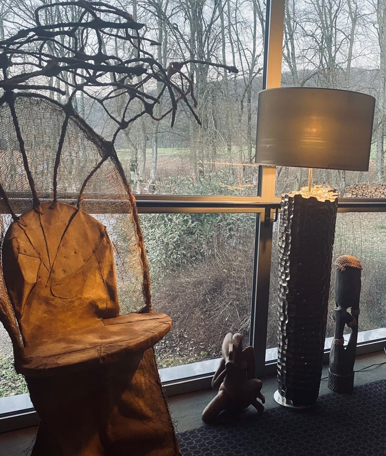 Een kamer met een groot raam, een stoel, een lamp en een klein tafeltje met een decoratief object.