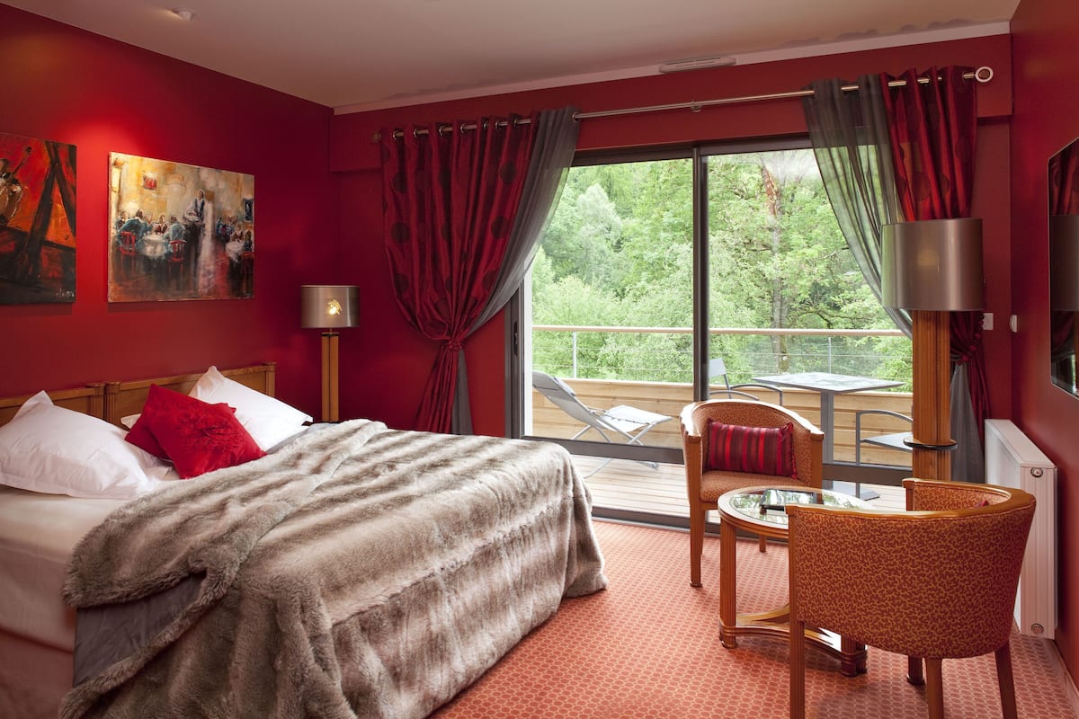 A hotel room with a bed, a seating area with a table and chairs, a painting on the wall, and a balcony with a view of greenery.