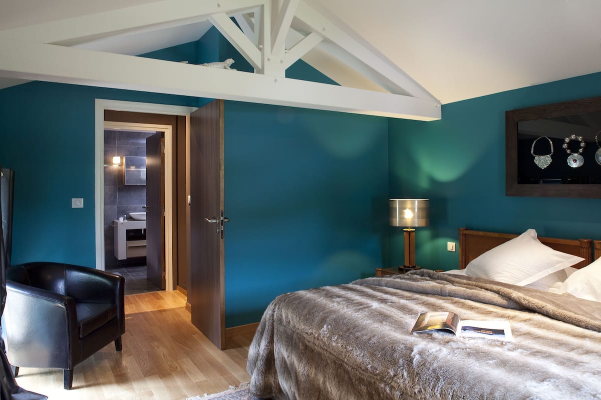 A bedroom with a large bed, a chair, a door leading to a bathroom, and a wall-mounted lamp.