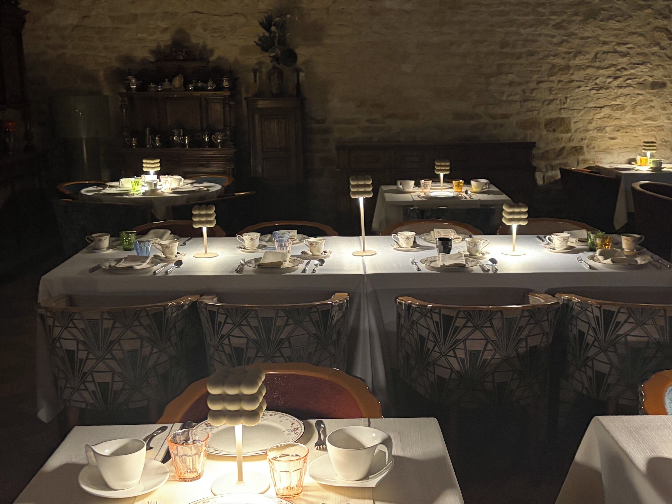 A dimly lit dining area with tables set for a meal, featuring a stone wall and wooden furniture.