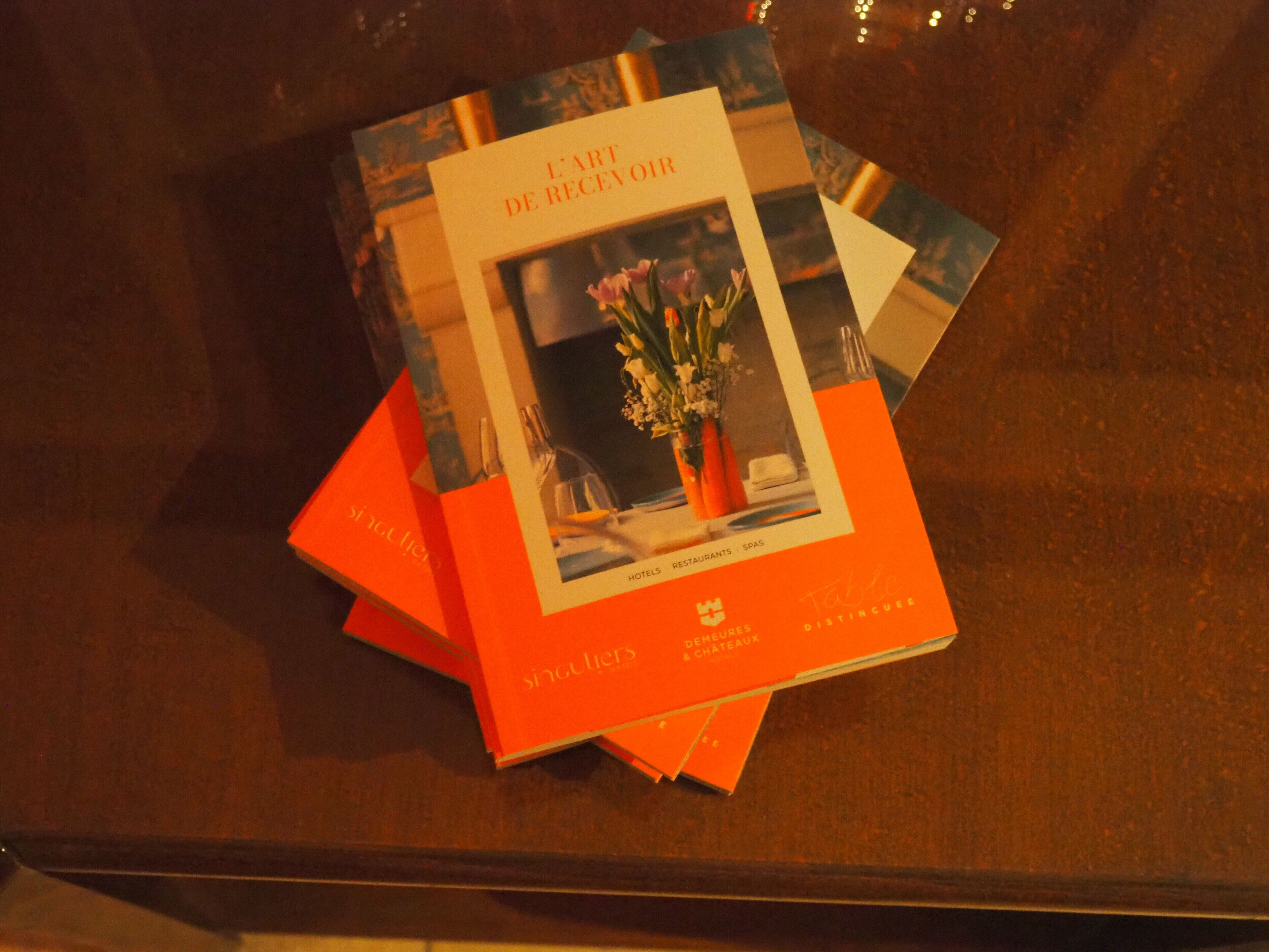 A brochure titled 'L'ART DE RECEVOIR' with a floral image on the cover, placed on a wooden surface.