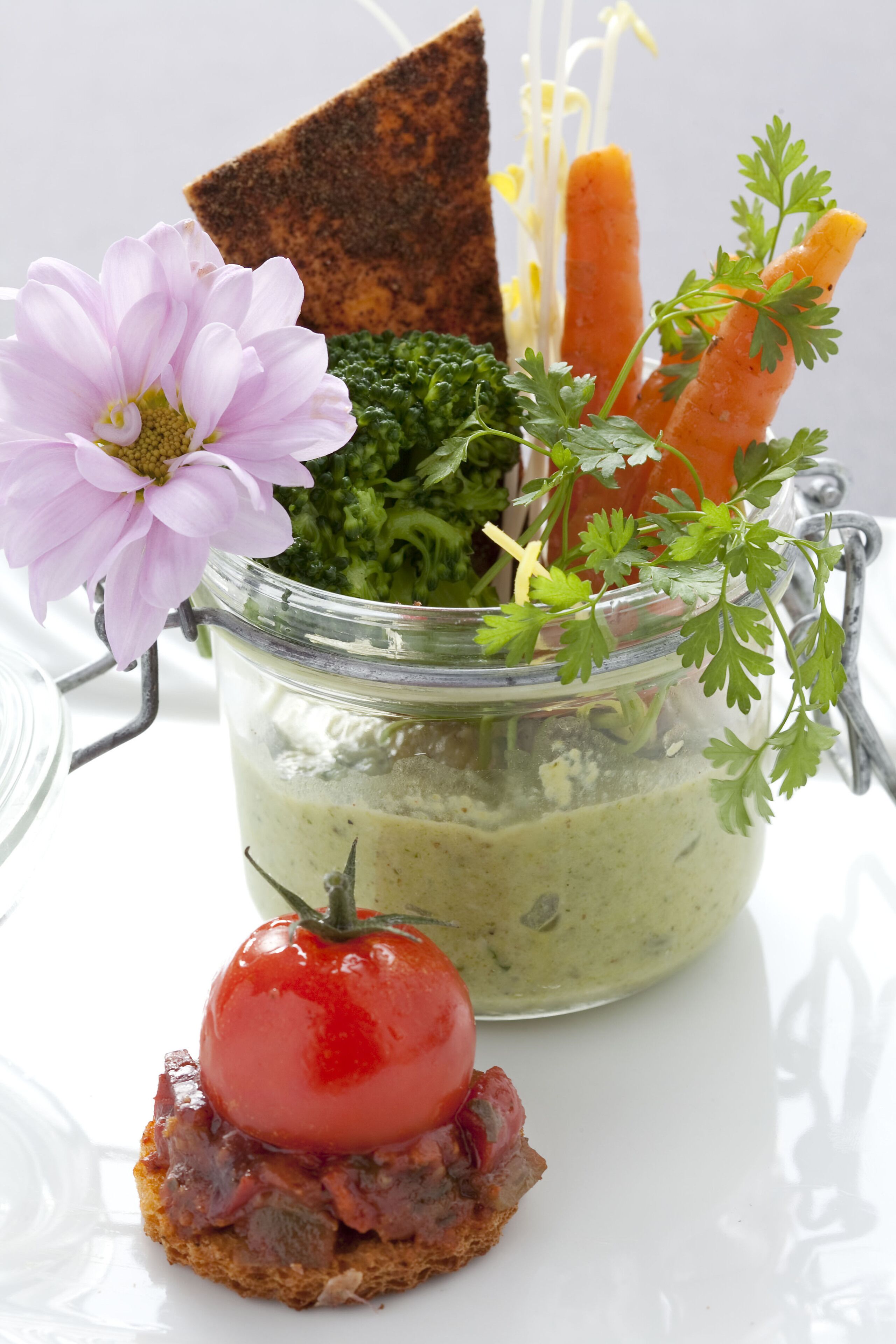 Ein Glas mit grüner Soße, einer Tomate und einem Stück Brot auf einer weißen Oberfläche.