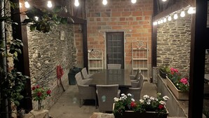 Une terrasse extérieure avec un mur en briques, des piliers en pierre, des meubles en osier et des guirlandes lumineuses.