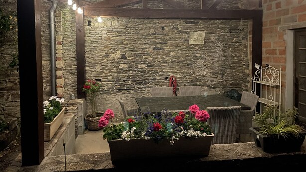 Un espace extérieur aménagé avec des murs en pierre, comprenant une table et des chaises, et entouré de plantes en pot et de guirlandes lumineuses.
