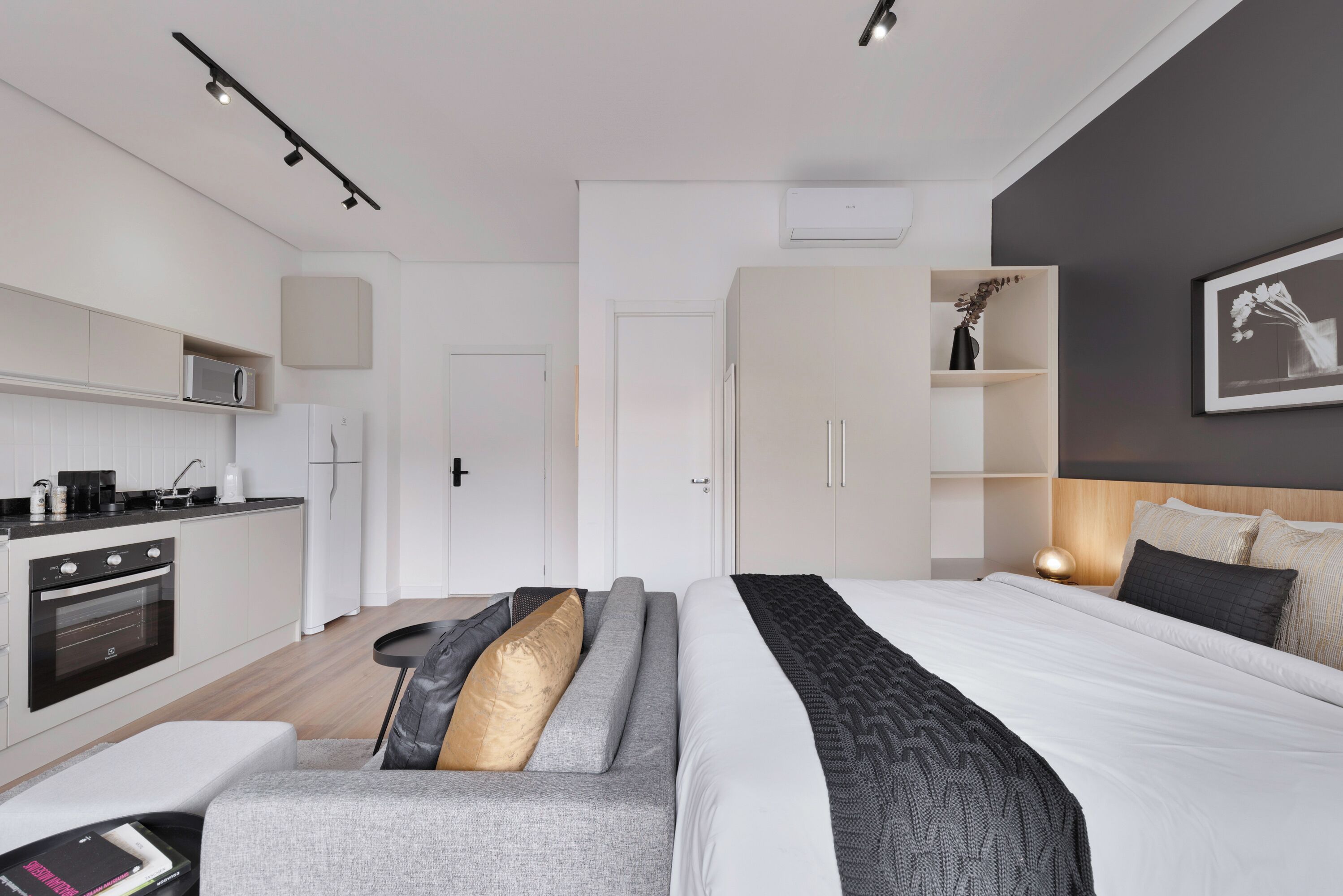 Ein modernes Hotelzimmer mit einer Küchenzeile, einem Sofa mit Kissen, einem Bett mit gemustertem Überwurf und einem Nachttisch mit Lampe.