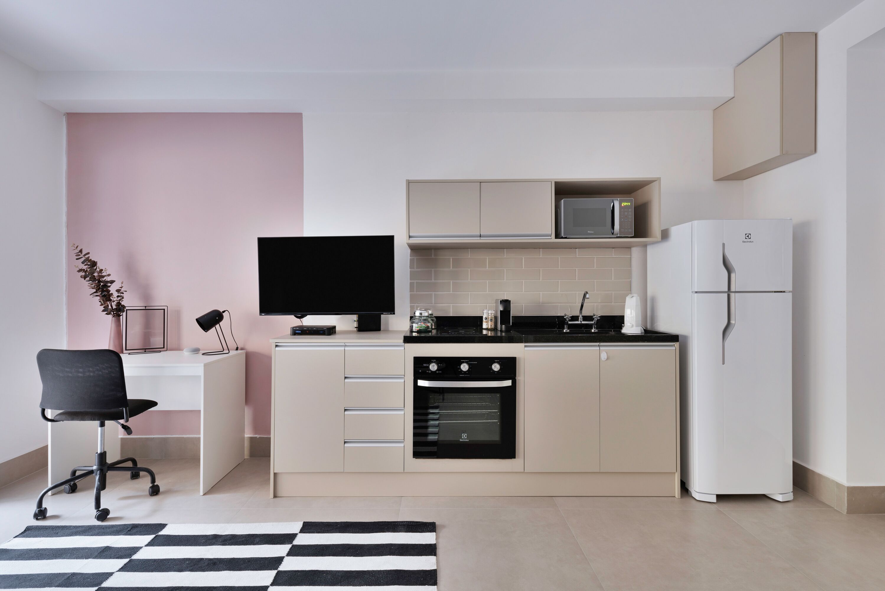 A modern kitchen with a built-in oven, microwave, and refrigerator. There is a black office chair and a flat-screen TV on a white desk.