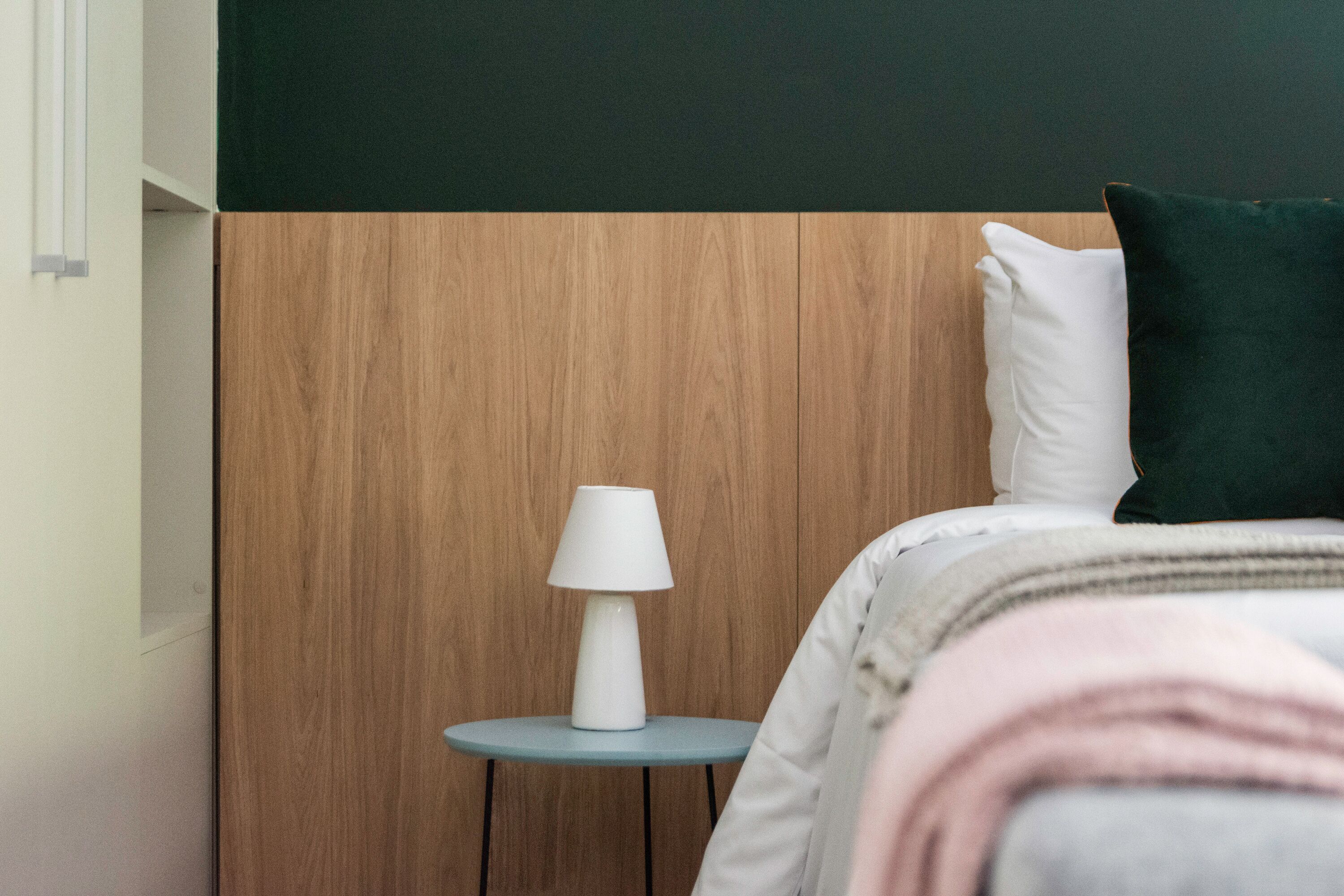 A neatly made bed with a white pillow and a dark green pillow, a white bedside lamp on a small blue table, and a wooden headboard.