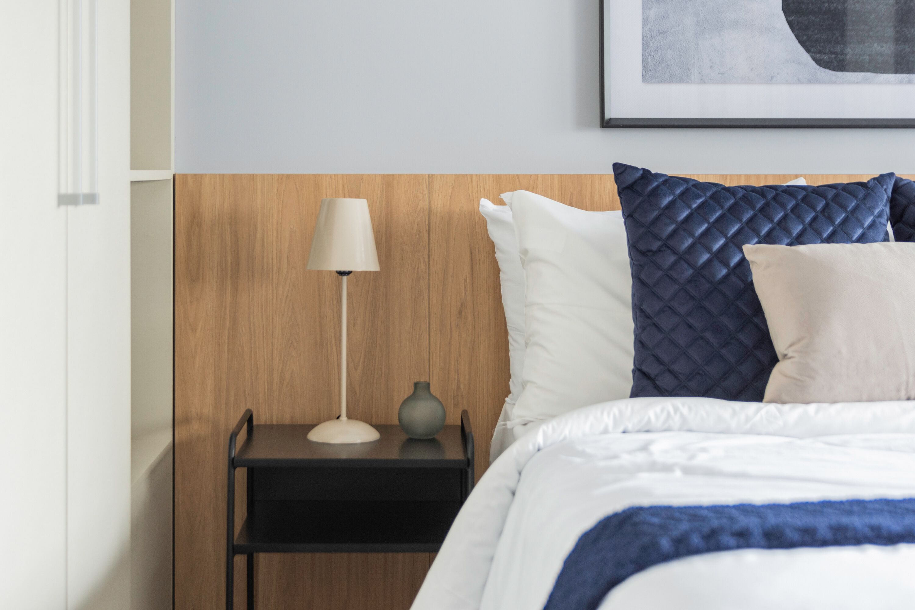 Uma cama bem arrumada com roupa de cama branca e azul, uma luminária de cabeceira e uma mesinha de noite com um vaso.