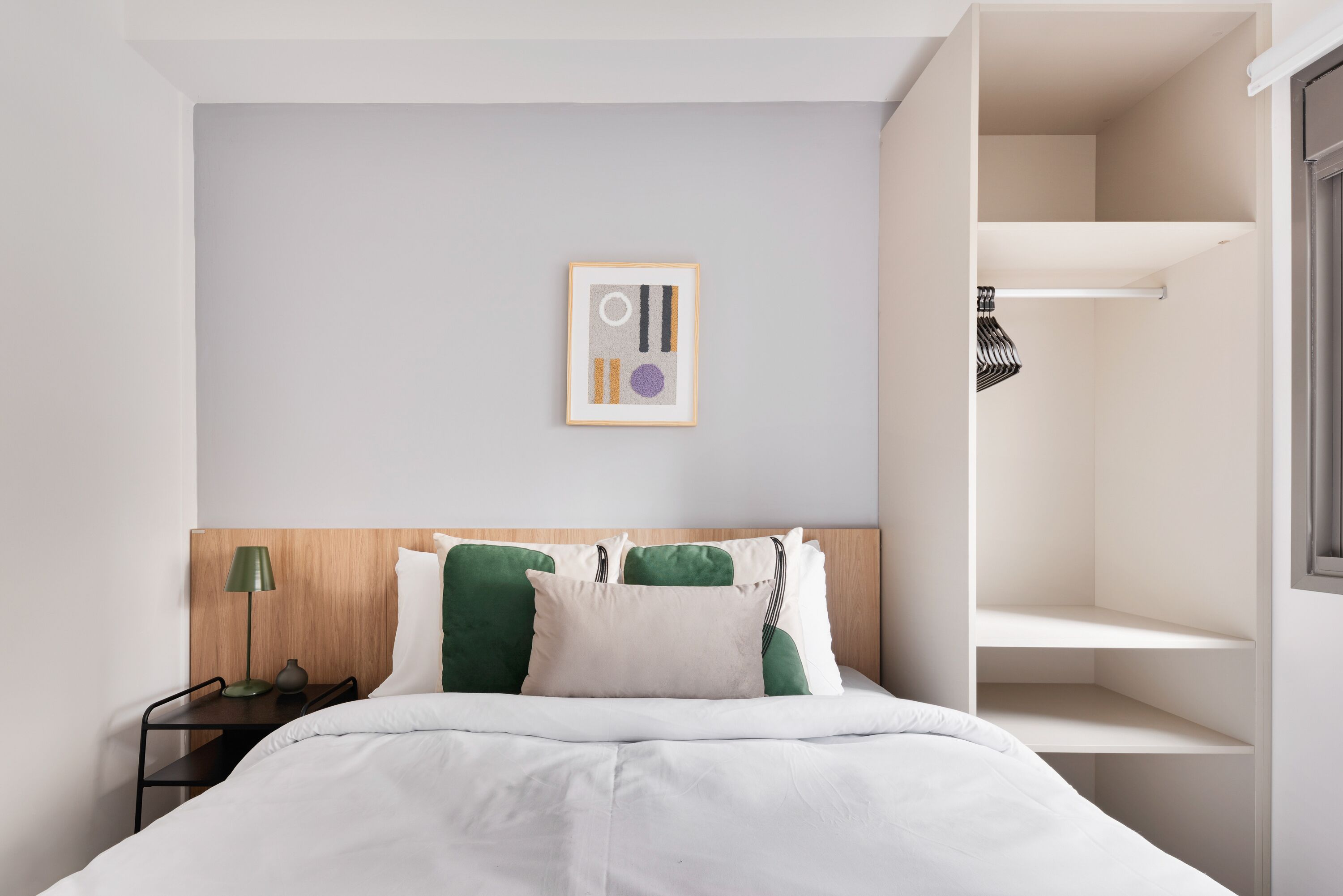 A neatly arranged hotel room with a bed, a bedside table with a lamp, a framed artwork on the wall, and a built-in closet with shelves.