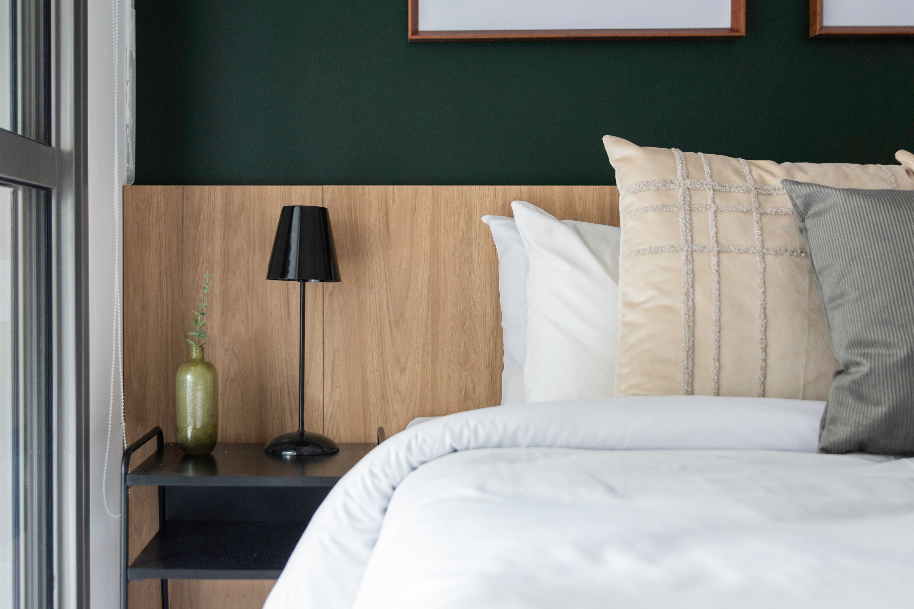 A neatly made bed with white and beige pillows, a black lamp on a wooden nightstand, and a green vase.