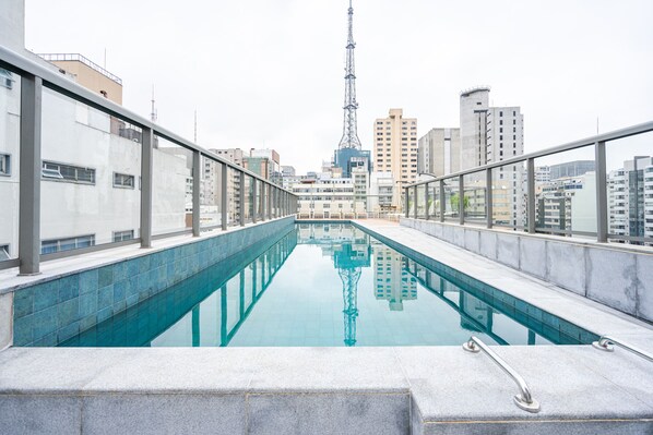 A rooftop swimming pool with a glass barrier, surrounded by modern buildings and a prominent tower in the background. - Charlie Edição Jardim Paulista (São Paulo)